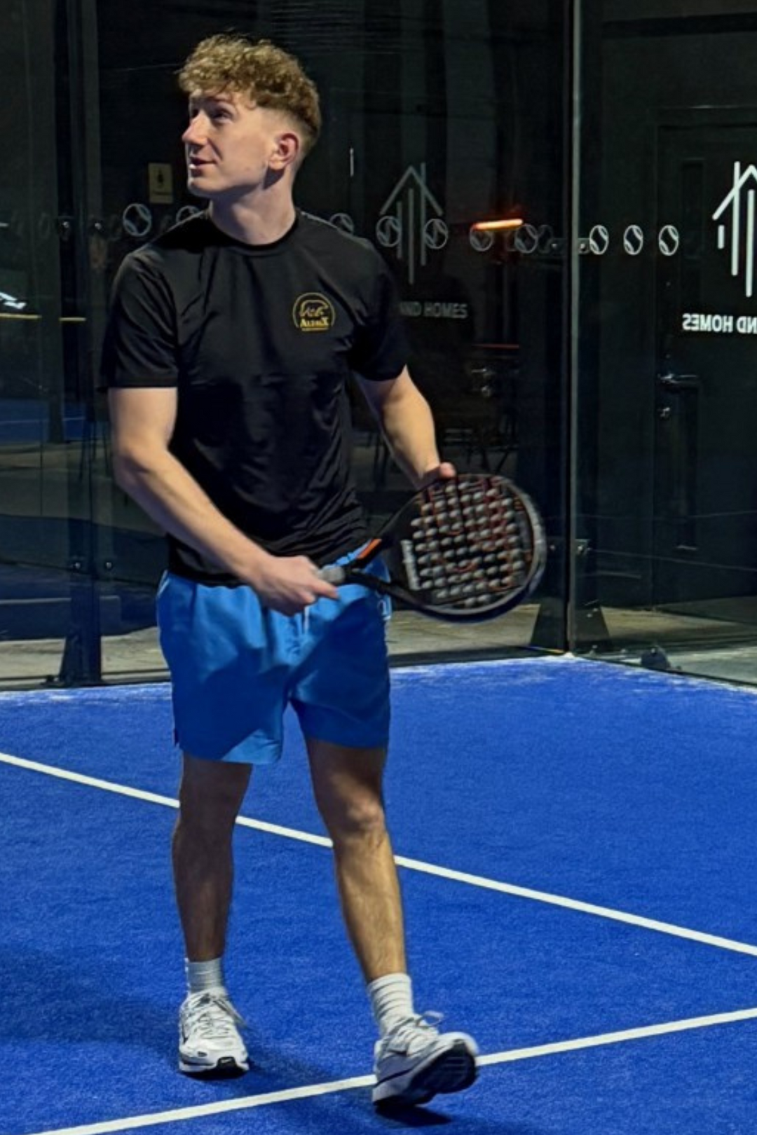 A man standing on a blue indoor court holding a paddle racket, wearing a black short-sleeve athletic shirt with a small gold chest logo, bright blue shorts, white socks, and white sneakers.