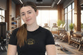 Woman in a gym wearing a black t-shirt with a logo, surrounded by fitness equipment.